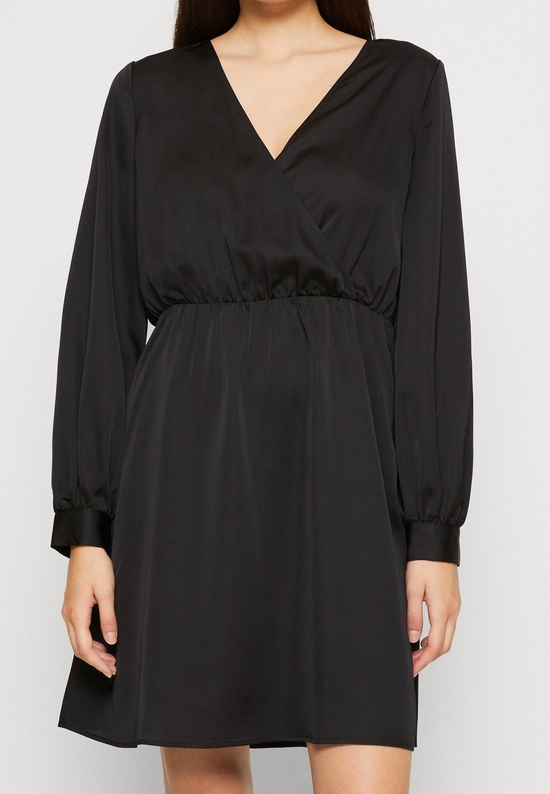 Femme portant une robe noire à manches longues avec un décolleté en V et une taille froncée, debout devant un fond clair uni.