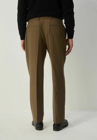 Henkilö, joka on pukeutunut ruskeisiin mittatilaus housuihin, mustiin kenkiin ja mustaan pitkähihaiseen puseroon, seisoo selkä kamerasta pois yksinkertaista vaaleaa taustaa vasten.