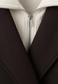 Chaqueta marrón con un cuello de canalé blanco. Cuenta con un cierre central con un tirador plateado y solapas angulares. Textura suave con un diseño estructurado.