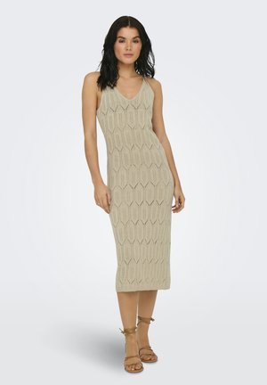 Mujer de cabello oscuro con un vestido midi de punto sin mangas de color beige claro y sandalias marrones con cordones, de pie frente a un fondo liso y claro.