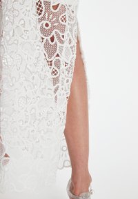 Weißes Spitzenkleid mit filigranen Blumenmustern und einem Oberschenkelspalt. Die Textur ist zart, mit transparenten Abschnitten und einer glatten Oberfläche.