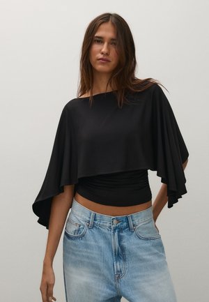 Une femme porte un haut noir drapé par-dessus un haut noir ajusté et un jean bleu clair, sur un fond uni.