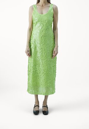 Femme debout portant une robe midi sans manches à sequins verts vifs avec des chaussures Mary Jane noires sur un fond blanc.