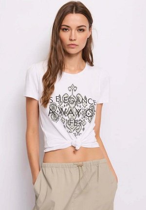 Giovane donna con lunghi capelli castani indossa una t-shirt bianca con nodo e pantaloni beige, in piedi con le mani nelle tasche davanti a uno sfondo semplice.