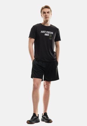 Maglietta sportiva nera con testo bianco e verde, abbinata a pantaloni corti neri con cuciture verde neon e scarpe da ginnastica nere.