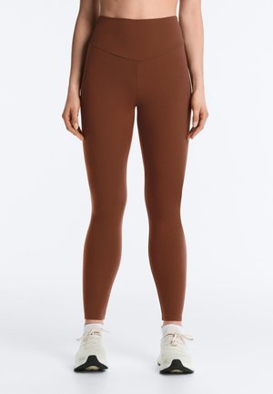 Nő, barna magasderekú leggingset és fehér tornacipőt visel, egy egyszerű világos háttér előtt áll.