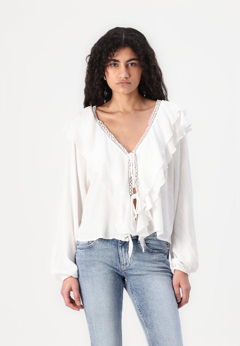 Blusa bianca con davanti a ruches, scollatura a V e chiusura con fiocco. Maniche lunghe e dettagli in pizzo delicato sul colletto. Tessuto texturizzato.
