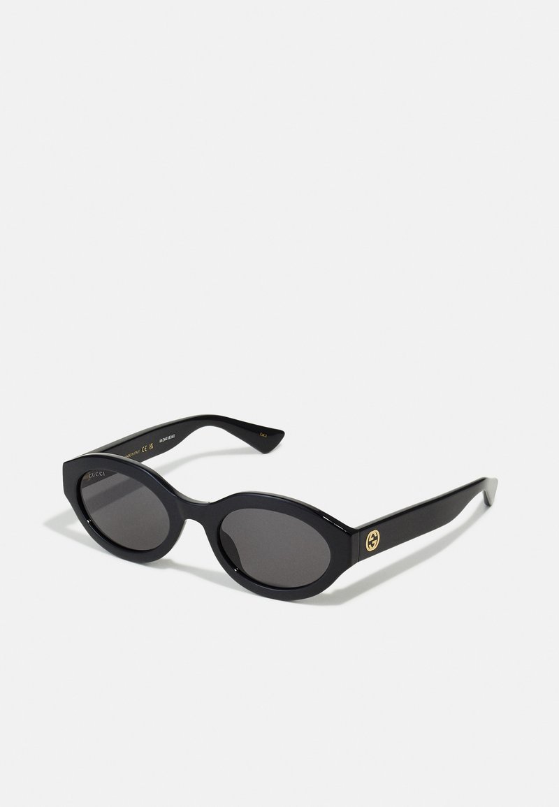 Óculos de sol ovais pretos com lentes escuras e armação de plástico brilhante. Apresenta um logotipo dourado na haste, oferecendo um design elegante e moderno.