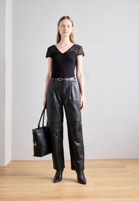Bodysuit noir avec des manches en dentelle, associé à un pantalon en cuir noir taille haute. Le modèle tient un sac fourre-tout noir et porte des bottes pointues.