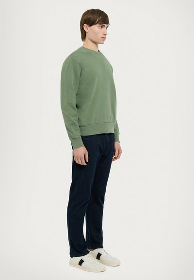 Felpa verde con girocollo, polsini e orlo a costine, abbinata a jeans blu scuro e scarpe da ginnastica bianche con dettagli neri.