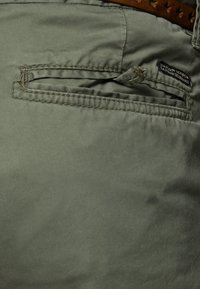 Pantaloni verde chiaro con una finitura testurizzata, dotati di una tasca laterale e una cintura in pelle marrone. Etichetta del marchio visibile vicino alla tasca.