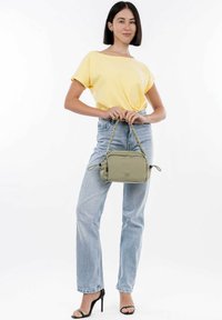 Borsa a tracolla in tessuto verde oliva con manico intrecciato, forma rettangolare e chiusura con zip. Il modello indossa un top giallo e jeans di un blu chiaro.