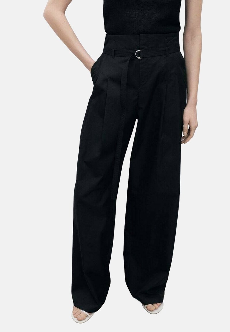 Femme portant un pantalon noir taille haute à jambes larges avec ceinture en tissu et un haut noir, debout avec une main dans la poche et des sandales blanches.