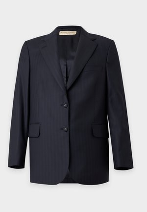 Femme portant un costume sombre oversize avec un pantalon à jambes larges, une chemise bleu clair boutonnée, des chaussures noires pointues et portant un petit sac à main noir.