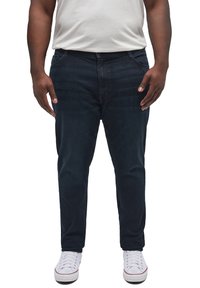 Bărbat purtând blugi albaștri închiși, adidași albi și un tricou alb, stând cu brațele relaxate pe lângă corp.