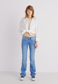 Camisa branca curta atada na cintura, blusa bege por baixo, jeans azuis boca de sino e tênis brancos, colocados contra um fundo simples.
