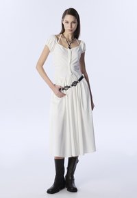 Vestido blanco de mangas cortas con cintura fruncida, cierre de cremallera y cinturón decorativo negro. Combinado con botas negras hasta la rodilla. Diseño minimalista.