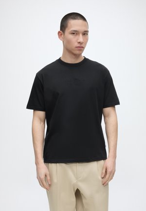 Mladý muž nosiaci čierne vyšívané tričko a béžové nohavice stojaci na jednofarebnom bielom pozadí.