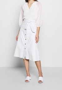 Blusa bianca trasparente con maniche a sbuffo e motivi sottili, abbinata a una gonna di jeans bianca a ginocchio con bottoni frontali e cintura annodata.