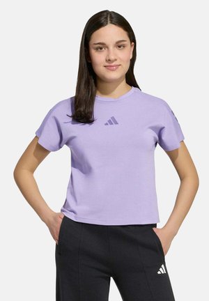 Junge Frau mit glatten dunklen Haaren, die ein hellviolettes Adidas-T-Shirt und schwarze Adidas-Hosen trägt und mit den Händen in den Taschen steht.