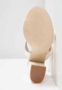 Sandale à talons blancs avec une tige en cuir lisse, bride de cheville réglable et un talon carré. Semelle en caoutchouc texturé pour une meilleure adhérence.