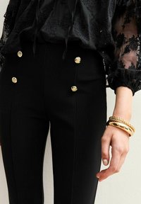 Pantalons noirs taille haute avec six boutons dorés associés à un chemisier en dentelle noire transparente et des bracelets en or au poignet.