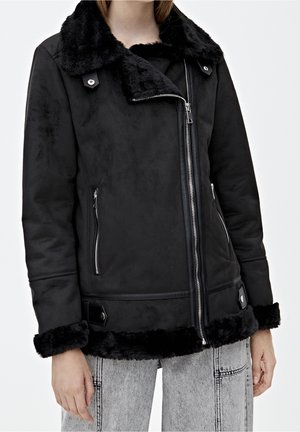 Veste noire doublée en fausse fourrure avec fermeture éclair argentée asymétrique, poches latérales zippées et détails à pattes à boutons-pression sur les poignets et l'ourlet.