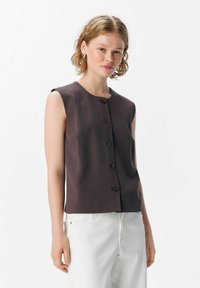 Haut sans manches marron avec un col rond et cinq boutons sur le devant. Texture lisse ; associé à un pantalon en denim blanc.