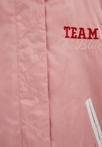 Rausvos spalvos pūsta striukės dalis su raudonu siuvinėtu užrašu „TEAM“ ir rožiniu užrašu „Blue“, taip pat vertikaliai baltu apvadais apjuostu kišenės plyšiu.