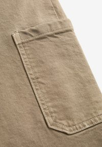 Poche en tissu khaki avec une bordure cousue, présentant un motif de couture diagonal et une surface texturée. La poche a une forme rectangulaire.