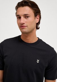 Zwarte katoenen t-shirt met een ronde hals en korte mouwen, met een klein wit chevron-logo op de linkerkant van de borst.