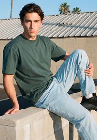 Noor mees rohelise t-särgiga ja helesinistes teksades istumas betoonist äärekivil õues, taustal on palmid ja selge taevas.