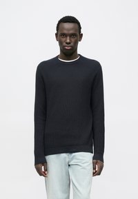 Pull en maille côtelée marine avec un col rond et des manches longues, associé à un jean bleu clair. Texture lisse, design minimaliste, sans motifs visibles.