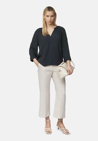 Zwarte blouse met lange mouwen en V-hals; beige, op maat gemaakte broek; witte sandalen met bandjes; kleine witte handtas met hengsels.