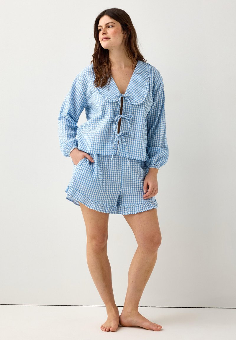 Γυναίκα που στέκεται ξυπόλητη φορώντας μπλε και λευκό καρό μακρυμάνικο τοπ με κορδόνια και ταιριαστό σορτς με βολάν, μπροστά από ένα απλό φόντο.