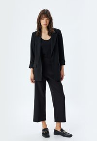 Zwarte op maat gemaakte blazer met opgerolde mouwen, gecombineerd met een zwart tanktop en brede broek. De schoeisel heeft glanzende, gestructureerde details.