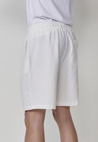 Persona che indossa pantaloni corti bianchi con tessuto testurizzato, dotati di vita elastica e tasche laterali, in piedi contro uno sfondo chiaro e uniforme.