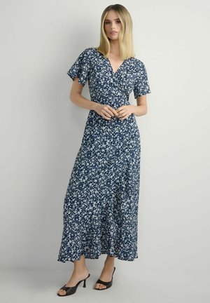 Kobieta w granatowej, kwiecistej sukience kopertowej z krótkimi rękawami i czarnych sandałach na obcasie stoi na tle jednolitego jasnego tła.