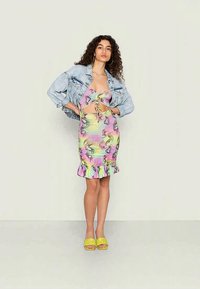 Mulher com cabelo encaracolado vestindo um vestido recortado em tie-dye pastel, uma jaqueta de denim clara e cropped, e sapatos de salto abertos amarelos, de pé contra um fundo simples.