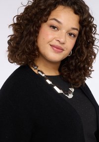 Cardigan nero con scollo a V e tessuto testurizzato, indossato sopra una maglietta nera con piccoli pois. Presenta una collana geometrica argento.