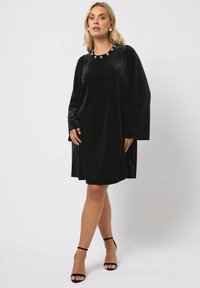 Schwarzes Samtkleid mit rundem Halsausschnitt, geschmückt mit dekorativen Verzierungen. Verfügt über lange Ärmel und eine lockere, fließende Silhouette.
