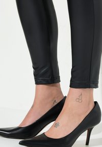 Leggings de cuero sintético negro combinados con elegantes tacones de punta negra. Los tatuajes visibles en los tobillos incluyen una flor y cerezas.