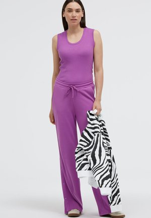 Frau steht und trägt ein lila ärmelloses Oberteil sowie passende Hosen mit Kordelzug, in der rechten Hand hält sie eine schwarz-weiß gestreifte Zebra-Jacke.