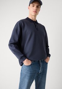 Granatowa koszula polo z długim rękawem, z kołnierzykiem i zapięciem na trzy guziki, zestawiona z jasnoniebieskimi dżinsami. Gładka faktura, luźny krój.
