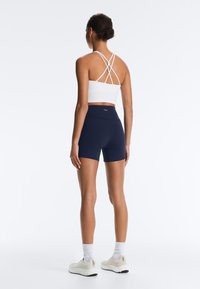 Nő áll háttal a képen, fehér pántos crop topot, tengerészkék magas derekú rövidnadrágot, fehér zoknit és bézs sportcipőt visel.