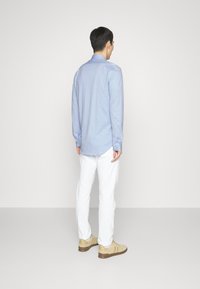 Vaaleansininen pitkähihainen paita, joka on valmistettu kudotusta kankaasta, yhdistetty valkoisiin housuihin ja beigen värisiin tennareihin. Näkyvät napit ja kaareva helma.