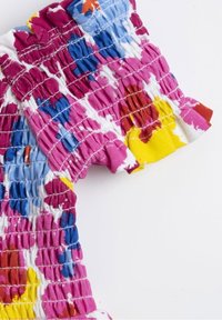 Top z smockované látky s živým květinovým vzorem v růžové, žluté, modré a červené; obsahuje volánkový výstřih a elastickou texturu.
