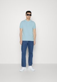 Jasnoniebieska koszulka polo z krótkimi rękawami i trzyguzikowym zapięciem, w połączeniu z niebieskimi dżinsami i białymi trampkami. Model w czarnych okularach przeciwsłonecznych.