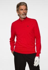 Homme âgé aux cheveux gris et à la barbe, portant une chemise rouge à manches longues, un pantalon noir et un gant blanc à la main droite, regardant sur le côté.