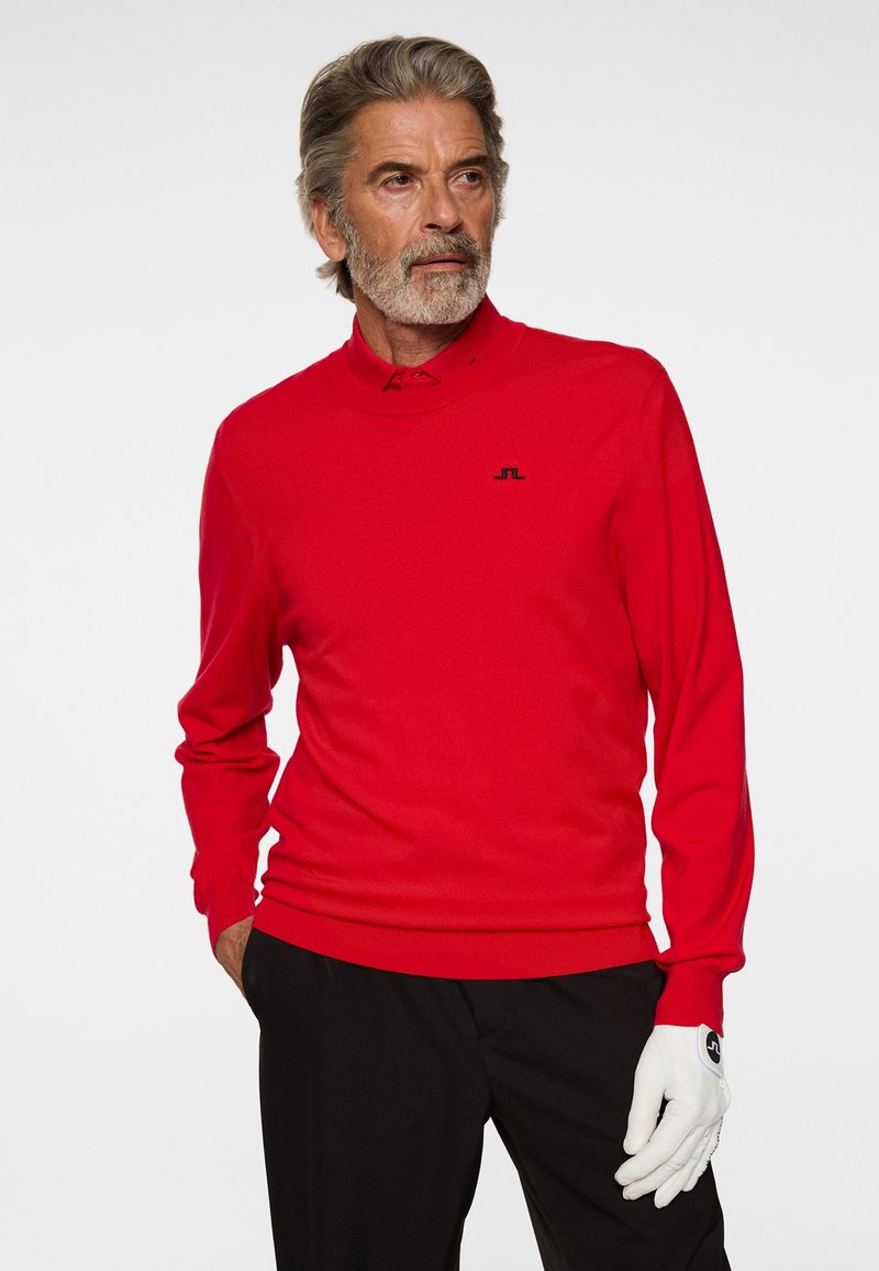 Älterer Mann mit grauen Haaren und Bart, der ein rotes Langarmshirt, schwarze Hose und einen weißen Handschuh an der rechten Hand trägt und zur Seite blickt.